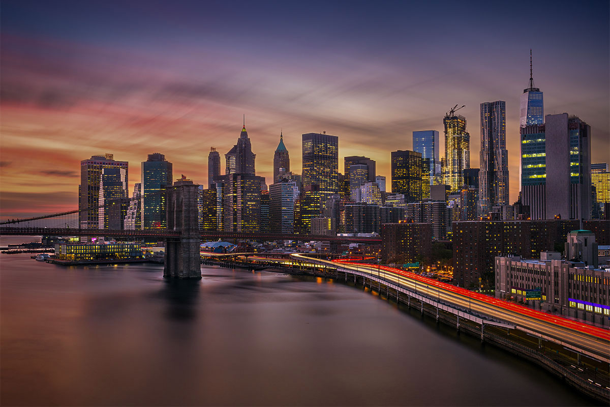 New York City skyline at sunset from the Manhattan Bridge, with light trails along the East River